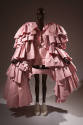 Pink harness with oversize layered sleeves of gathered flounces reaching to floor over matching…