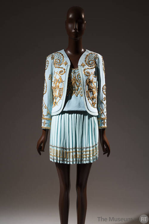 Light blue suit with allover gold tone studs in spiral patters on collarless jacket and Greek k…
