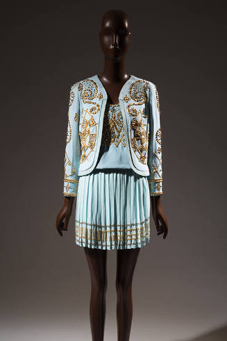 Light blue suit with allover gold tone studs in spiral patters on collarless jacket and Greek k…