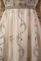 Detail view of white muslin dress showing metallic braided trim around empire waist and metalli…