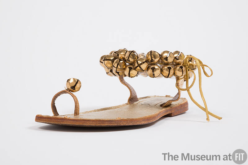 Flat leather sandal with ankle strap covered in gold bells and single big toe loop affixed with…