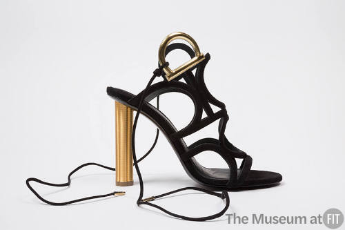 Black sandal with thin straps of linking circles, tie ankle straps attached by gold hardware an…