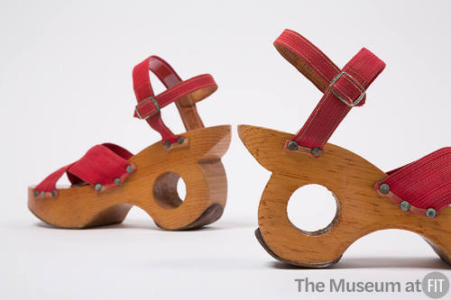 Pair of sandals with red elastic straps crossed at vamp and attached to wooden heel with silver…