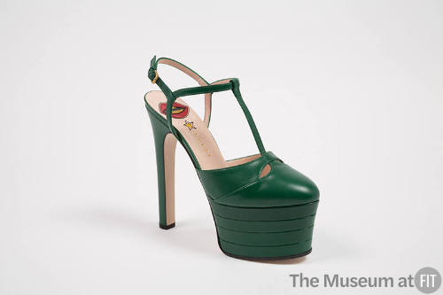 Green high heel platform t-strap shoe with Gucci logo, yellow star and red mouth visible on bei…
