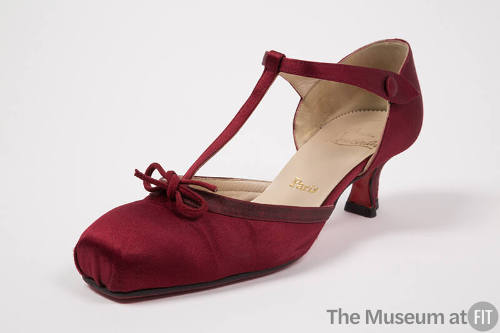 Dark red T-strap shoe with square toe box, small bow at vamp, low heel and gold “Paris” visible…