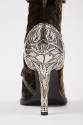 Detail view of brown velvet ankle boot showing silver stiletto heel with a bird, vine, heart an…