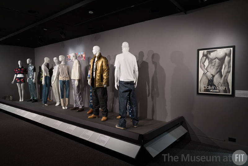 Denim_07 Left to right 86.151.1 (jacket and skirt), 2012.19.1 (beaded ensemble), 2013.36.2 (jacket hidden), 2015.63.1 (jeans), 90.150.20 (light jeans), 2015.51.9 and 2015.51.8 (top and jeans), 91.256.2 and 2010.74.29 (sweater and jeans), 92.10.2 (ensemble hidden),  99.122.2 (gold coat ensemble), 2013.62.6 and 2013.62.8 (ensemble hidden), 2000.86.1 (jeans), 93.58.78 (poster)