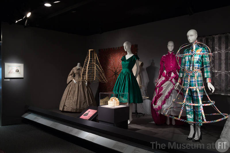 RetroSpective_04 left to right P88.14.5 (dress), 86.157.9 (shawl wall), 84.26.2 (hoop hanging), 2001.73.1 (crinoline), 92.59.1 (green dress), 2001.73.1B (hoop package),  P91.38.4 (hoops case), 2007.33.2 (purple and white dress), 2013.20.1 (ensemble), 91.187.21 (textile wall) 
