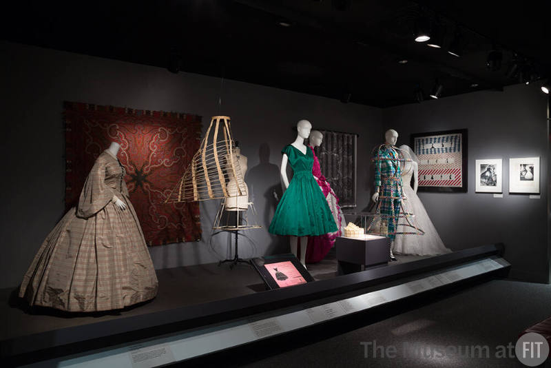 RetroSpective_32 Left to right P88.14.5 (dress), 86.157.9 (shawl wall), 84.26.2 (hoop hanging), 2001.73.1 (crinoline), 92.59.1 (green dress), 2001.73.1B (hoop package),  P91.38.4 (hoops case), 2007.33.2 (purple and white dress), 2013.20.1 (ensemble), 91.187.21 (textile wall), 2007.53.1 (wedding gown), 94.5.1 (scarf side wall)