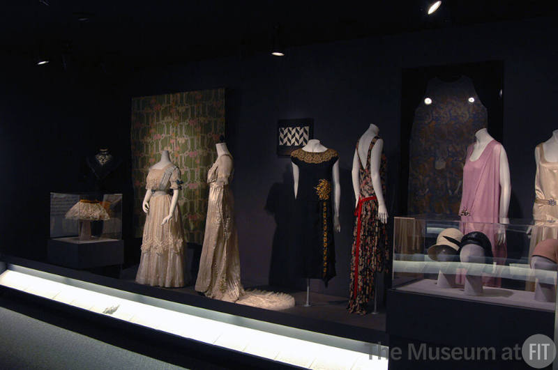 Arbiters_59 Left to right 72.66.6 (dress), P83.19.12 (hat case), P92.34.6 (textile wall), 95.145.13 (lace and satin dress), 95.145.10 (wedding dress), X321.7A (textile wall), P91.76.1 (black dress), 71.268.7 (floral dress), P89.25.1A & F (textile wall), 84.201.1 (pink dress), 83.219.6 (dress), P83.39.14, P83.39.16, 77.178.10 (hats case)