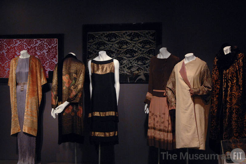 Exoticism_11 Left to right 83.151.1 (gown), 2007.22.1 (velvet coat), 73.65.5 (evening coat), 2002.87.1 (black dress), 83.79.2421 (textile wall), 83.79.49 (textile wall), P76.1.1 (dress), 84.213.1 (off white coat), 77.225.2 (cape)