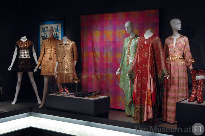 Exoticism_23 Left to right P88.1.1 (mini dress), 86.15.6 (scarf wall), 2002.58.1 (gold dress), 76.185.3 (gold suit), 93.148.10 (shoes case), U.2183 (earrings case), 86.104.2 (hat case), 68.143.94 (bag case), 76.56.16 (shoes case), 94.85.2A (textile wall), 86.130.61 (green ensemble), 2007.46.24 (caftan), 80.58.5 (pink evening dress), 97.60.40 (boots case)