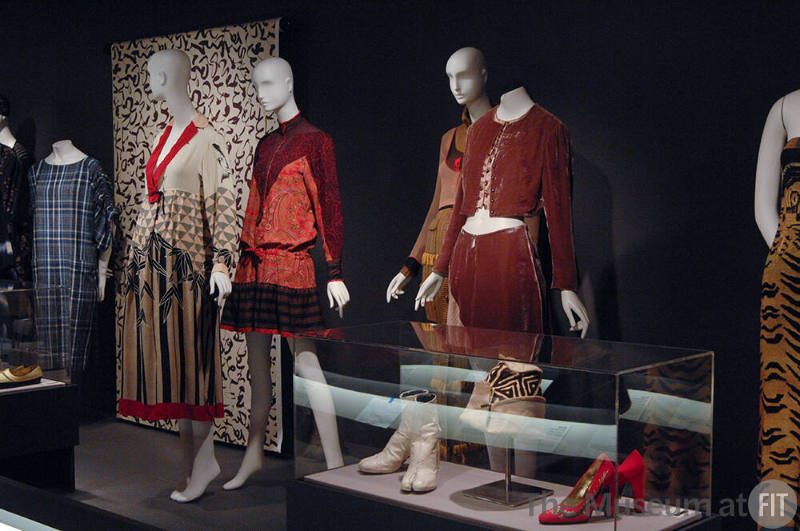 Exoticism_27 Left to right 2003.28.2 (plaid dress), 92.229.9 (textile wall), 96.6.23 (dress), 87.64.1 (paisley dress), 95.7.2 (sweater ensemble hidden), 99.53.1 (pink velvet suit), 92.182.1 (boots case), 93.165.2 (hat case), 98.13.1 (shoes case), 98.50.1 (tiger print dress partial)