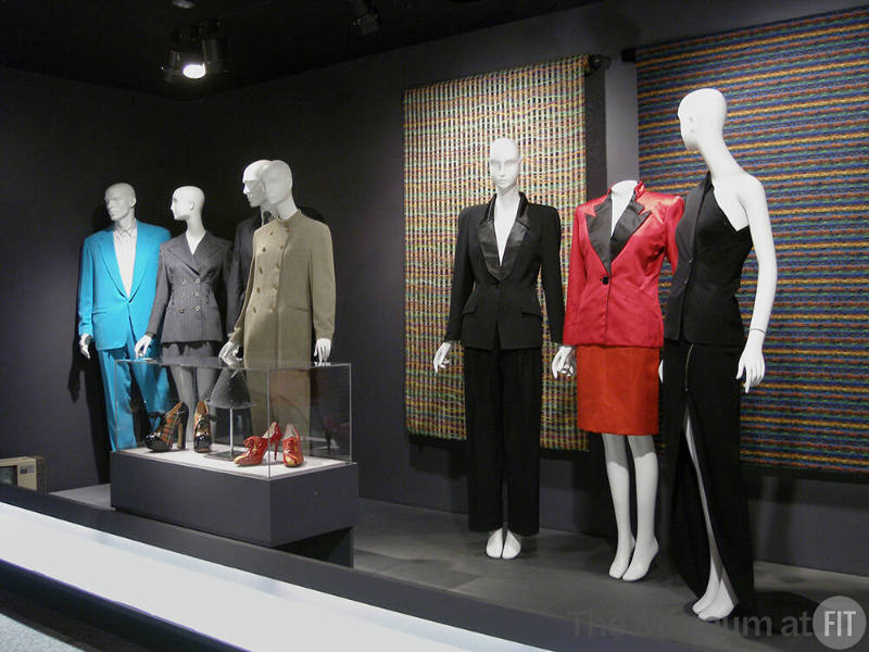 Tailor_14 Left to right  98.20.11 (blue suit), 95.77.2 (grey shorts suit), 2003.109.1 (dark blue suit hidden), 2006.7.4 (tan pantsuit), 98.119.1 and 99.3.1 (shoes case), 95.83.1 (hat case), 2005.90.9 (black tuxedo), 2005.90.6 (red suit), 97.71.1 (ensemble), 2006.11.4 and 2006.11.3 (textiles wall)