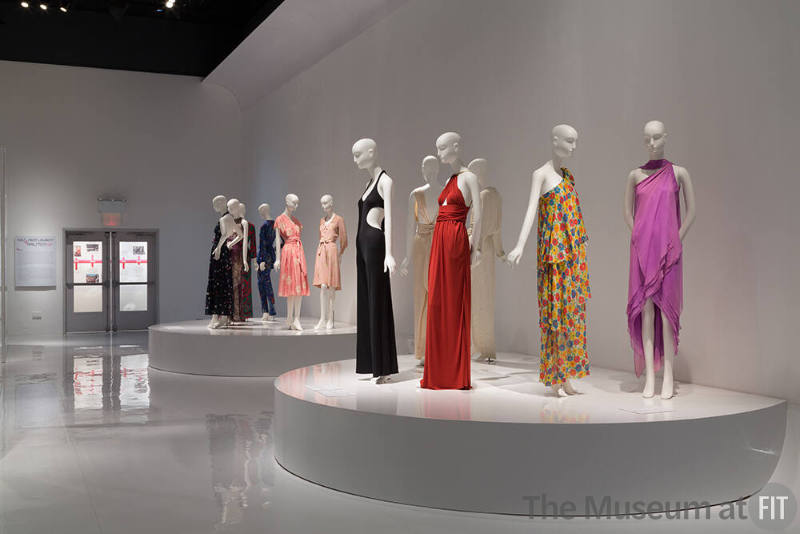 YSL+Halston_25 Left to right  91.41.1 (floral dress), 86.126.14 (purple grey dress), P89.55.4 (paisley ensemble), 91.41.12 (blue ensemble), 2014.28.1 (pink dress), 93.1.26 (top and skirt), 91.104.1 (white dress), 91.235.4 (halter dress),  79.66.40 (black jumpsuit), 88.1.69 (orange dress), 87.146.27 (floral dress), 82.3.5 (purple dress)