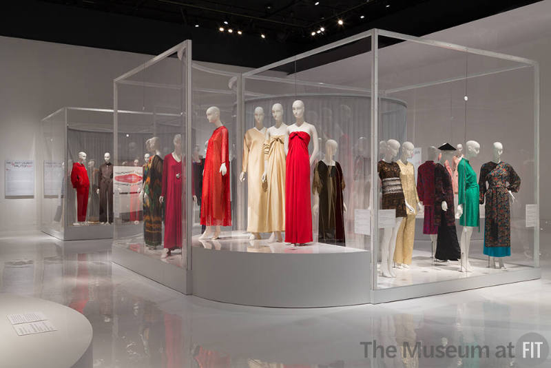 YSL+Halston_31 Left to right 96.119.11 (red jacket and pants), 86.126.11 (jumpsuit), 91.241.11 (brown tie dye ensemble), 76.69.8 (red dress),  82.3.1 (orange caftan), 92.109.4 (satin dress), 82.3.9 (dress and stole), 80.128.4 (red dress), 88.81.1 (black dress), P88.1.1 (beaded dress), 88.178.1 (gold pantsuit), 92.222.7 (green dress), 93.98.1 (hat), 2006.35.2 (dress), 86.126.13C (black pants), 88.73.1 (printed silk ensemble)