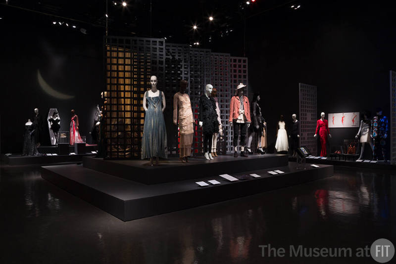 Exhibitionism_40 Left to right  2007.23.2 (beaded dress), 2007.2.1 (black ensemble),  99.80.1 (velvet dress), 2008.5.1 (hat case), 2018.3.2 (earrings case), 2008.55.1 (gown with red), 2010.37.12 (denim dress), 2009.62.1 (dress), 2010.57.1 (black ensemble), 2009.75.1 (coat enemble hidden), 2009.61.1 (pink jacket ensemble), 2008.57.1 (black ensemble with hat), 72.61.91 (white dress), 88.157.3 (top and skirt), 71.265.13 (red dress), 87.16.1 (shoes case), P86.52.1 (bag case), 86.10.1 (paper dress), P85.29.1 (suit)
