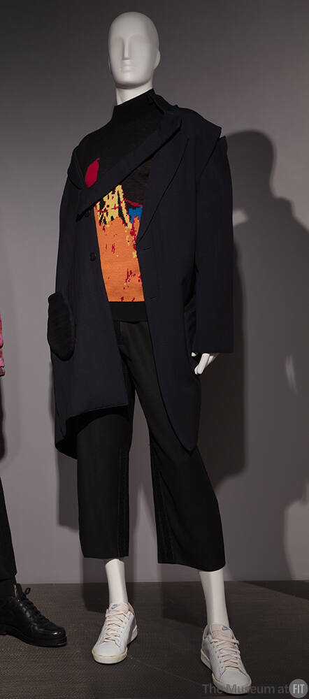 Man’s navy blue coat with diagonal opening and lapel panel over black sweater with orange abstr…