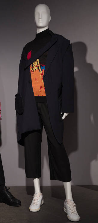 Man’s navy blue coat with diagonal opening and lapel panel over black sweater with orange abstr…