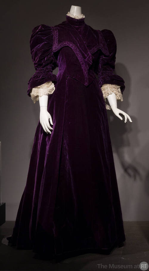 Purple floor-length gown with high neck and 3/4-length puffed sleeves, cinched at the elbow wit…